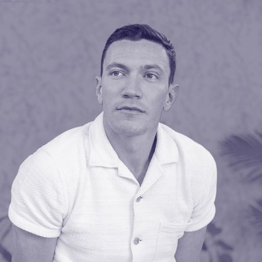 David Perell, in a short-sleeve white shirt, sits in front of a blurred background with plants, looking slightly to the side.